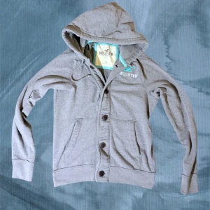 Grå hoodie från Hollister med knappar - Snygg grå hoodie från Hollister med knappar framtill och två fickor. Tröjan har huva med snörning och turkosa detaljer vid loggan och insidan av huvan. Perfekt för chill dagar och enkel att matcha med jeans eller joggers.