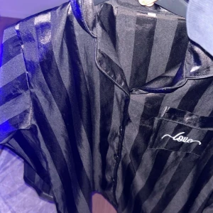 Svart randig satin-skjorta med broderi med byxor  - 300 för tröjan 450 för tröjan med matchande byxor. svart skjorta  i glansigt satinmaterial med breda ränder och klassisk krage. På bröstfickan finns ett broderat 'love'-motiv i vitt. Skjortan har knappar framtill och en loose passform som ger en chill vibe.