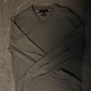 Grå v-ringad tröja Tommy Hilfiger - Säljer en stilren grå v-ringad tröja från Tommy Hilfiger med diskret logga på bröstet. Tröjan har lång ärm, ribbade muddar och är gjord i mjuk bomull blandad med cashmere. Perfekt för dig som gillar klassisk och enkel stil.