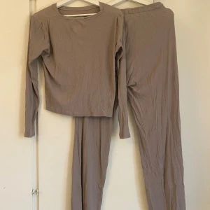 Brun/beige ribbstickat set - En basic beige långärmad tröja i ribbad bomullsmix och ett par byxor i samma material. Tröjan har rund halsringning och en rak, något lös passform. Byxorna sitter löst och skönt på benen. Materialet är lite urtvättat men passar perfekt till en kväll hemma eller liknande