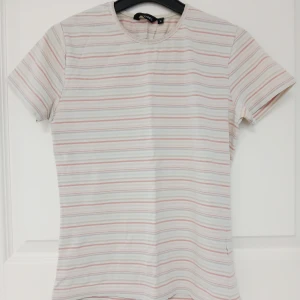 Randig t-shirt med korta ärmar - T-shirt med tunna horisontella ränder. Modellen har en rund halsringning och korta ärmar, perfekt för en chill look. Tillverkad i mjuk bomull (95 %) och elastisk elastan (5 %). Passar dig som gillar stilrena och ljusa färger. Plagget har naturligtvis tvättats. Se bilder för skick och passform. Storlek M enligt lappen men då måttet armhåla till armhåla är cirka 41 cm så stämmer S bättre. Axelsöm till ärmslut är cirka 15-16 cm. Tvättas i 40 grader.