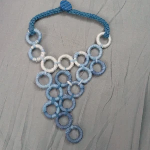 Handgjort blått halsband i garn - Unikt halsband med stora cirklar i olika blå och vita nyanser, inslagna i garn. Halsbandet har en flätad rem och knäpps med en blå knapp. Statement-piece som verkligen sticker ut med sin kreativa design och coola färgskiftningar.