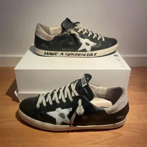 Golden Goose svarta sneakers (sällsynta) - Svarta Golden Goose sneakers i skinn med vit stjärndetalj på sidan, grå mockadetaljer vid hälen och vita snören. Sulan har texten 'Have a Golden Day' 