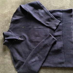 Mörkblå stickad tröja från Polo Ralph Lauren - Mörkblå stickad tröja från Polo Ralph Lauren med ribbad krage och två knappar vid halsen. Klassisk grön broderad logga på bröstet och breda ribbade muddar vid ärmslut och nederkant. Perfekt för lager-på-lager och chill stil.