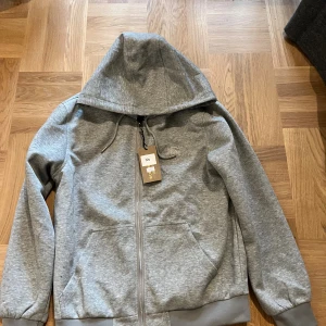 Grå hoodie från Burberry, XS - Snygg grå hoodie från Burberry i storlek XS. Tröjan har dragkedja, huva med snörning och broderad logga på bröstet. Tillverkad i mjuk bomullsmix och har ribbade muddar samt fickor framtill. Perfekt för en chill och stilren look.