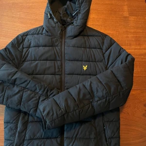 Svart pufferjacka från Lyle & Scott - Svart pufferjacka från Lyle & Scott med huva och gul broderad logga på bröstet. Jackan har quiltad design, dragkedja framtill och justerbara snören vid huvan. Perfekt för kyliga dagar och har en clean, modern look.