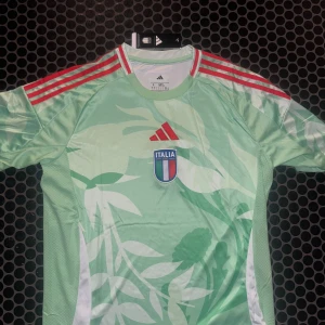 Italien Adidas fotbollströja mintgrön L - Italiens fotbollströja från Adidas i mintgrönt med vita och gröna bladmönster, röda Adidas-ränder på axlarna och broderad logga. Tröjan har korta ärmar och är tillverkad i ett lätt, ventilerande funktionsmaterial. Perfekt för dig som vill sticka ut på planen. Bara höra av er vid frågor/funderingar Fraktar samma dag som köp🚚
