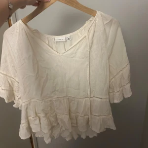 Vit volangblus från Vila - Söt vit blus från Vila med volangdetaljer och v-ringning. Blusen har korta ärmar, knytband vid halsen och fina spetsinslag längs volangerna. Perfekt för en luftig och romantisk look.
