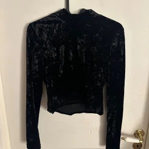 Svart sammet polotopp från Gina Tricot - Säljer en svart polotopp i mjuk sammet från Gina Tricot, storlek S. Toppen har lång ärm och croppad passform med en snygg glansig yta som ger extra attityd. Perfekt för dig som gillar stilrena och trendiga plagg med lite edge.