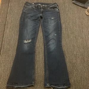 Lågmidjade jeans med hål - Snygga mörkblå jeans med bootcut passform och slitna detaljer på låret och knät. Jeansen har klassisk femficksdesign, dragkedja och knapp framtill. Materialet är jeans med en skön stretch och tvättad look som ger en cool vibe.
