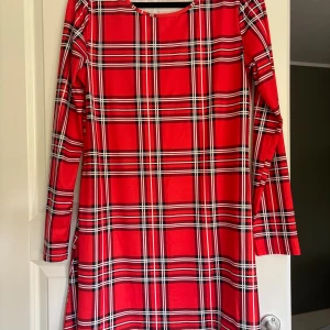 Röd rutig långärmad klänning - Snygg röd klänning med klassiskt rutigt mönster i svart och vitt. Modellen har långa ärmar och rak passform, perfekt för dig som gillar färgstarka plagg med retrovibbar. Materialet känns mjukt och följsamt mot huden.
