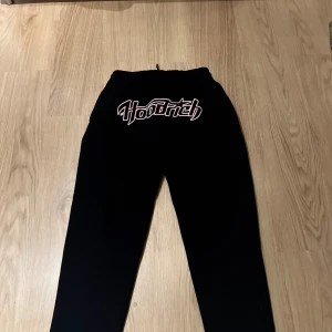 Svarta Hoodrich mjukisbyxor med broderi - Svarta mjukisbyxor från Hoodrich med stort broderat Hoodrich-logo i vitt och rött framtill. Byxorna har snörning i midjan och är tillverkade i mjukt material som är perfekt för chill eller streetwear. Passformen är loose och midjan är midwaist. 