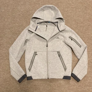 Ljusgrå hoodie från J.Lindeberg - Snygg ljusgrå hoodie från J.Lindeberg med hel dragkedja, huva och tre fickor med dragkedja. Mörkgrå detaljer vid ärmslut och fickor. Perfekt för lager-på-lager och chill dagar. Diskret logga på bröstet. Skick 8/10 använd fåtalet gånger och inga defekter, hör av er om frågor!