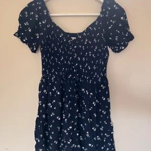 Blommig marinblå kortklänning/jumper Hollister - Supersöt marinblå kortklänning/jumper från Hollister med små vita blommor, puffiga korta ärmar och smockad överdel. Klänningen har volangdetaljer och en lätt, flowy kjol. Perfekt för sommarens alla tillfällen!