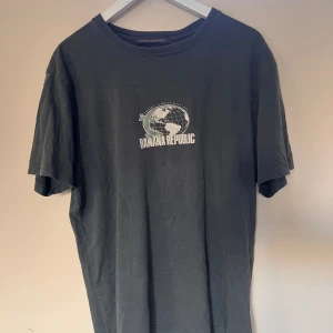 Grön/grå  t-shirt från Banana Republic - Svart t-shirt från Banana Republic med tryckt logga och jordglob på bröstet. Klassisk rund halsringning och normal passform. Tillverkad i 100% bomull för en skön och avslappnad känsla.