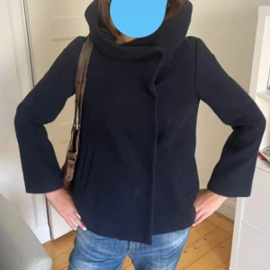 Mörkblå kappa  med stor krage - Snygg mörkblå kappa med unik, stor krage som ger en cool och modern look. Jackan har raka ärmar och en stilren, minimalistisk design utan synliga knappar. Perfekt för dig som gillar en clean och trendig stil. Den har en liten fläck men går nog bort om man tvättar hår själv inte provat. 