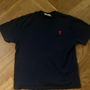 Svart t-shirt från Ami Paris - Svart t-shirt från Ami Paris i storlek S. Klassisk modell med kort ärm och rund halsringning. På bröstet finns Ami:s ikoniska röda hjärta-broderi. T-shirten är i mjuk bomull och har en clean, stilren look.
