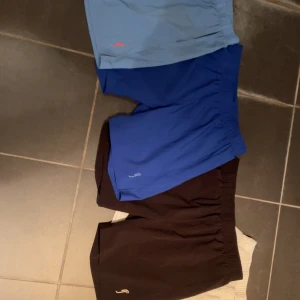 Färgglada träningsshorts i olika färger✨ - Fyra par shorts i blått, ljusblått, svart och vitt med elastisk midja och liten logga framtill. Perfekta för träning eller chill, med enkel och sportig design. Materialet känns lätt och smidigt, vilket gör dem sköna att bära under varma dagar. | passar killar som är ca 150-165,det beror på hur man gillar dem. | För mer frågor och funderingar skriv till mig.✨