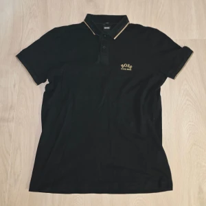BOSS Piké - Svart pikétröja från BOSS med gulddetaljer på krage och ärmslut. Slim fit-modell med klassisk krage och knappar framtill. Broderad BOSS-logga i gult på bröstet. Tillverkad i mjuk bomull för en skön känsla.