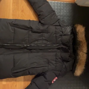 Svart dunjacka från Canada Goose S - Säljer en svart dunjacka från Canada Goose med klassisk patch på ärmen och stor huva med äkta päls. Jackan har quiltad design, dragkedja framtill och flera fickor. Perfekt för kalla vinterdagar och riktigt snygg streetstil.