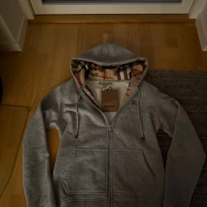 Grå hoodie från Burberry med dragkedja - Snygg grå hoodie från Burberry med klassiskt rutigt foder i huvan och broderad logga på bröstet. Tröjan har hel dragkedja, snörning i huvan och två fickor framtill. Perfekt för dig som gillar stilrena och exklusiva plagg.