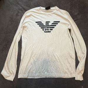 Vit långärmad tröja Emporio Armani - Vit långärmad tröja från Emporio Armani med stort svart logotryck på bröstet. Tröjan har en klassisk rund halsringning och är tillverkad i mjuk bomull, perfekt för en clean och stilren look.