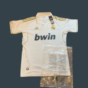 Real Madrid 11/12 Ronaldo fotbollströja - Vit Real Madrid fotbollströja från Adidas med guldiga detaljer och Bwin-tryck på bröstet. Nummer 7 och Ronaldo på ryggen i guld. Klassisk krage och korta ärmar. Materialet är lätt och andas, perfekt för match eller träning.