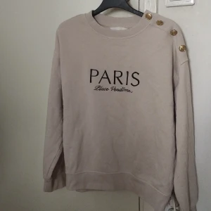 Beige sweatshirt med guldknappar H&M - Beige sweatshirt från H&M med svart text 'PARIS Place Vendôme' framtill. Tröjan har rund hals, långa ärmar och tre dekorativa guldknappar på ena axeln. Mjuk insida och relaxed fit, perfekt för en chill look.