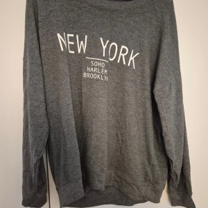 Grå sweatshirt New York Gina Tricot - Mysig grå sweatshirt från Gina Tricot med vit text 'New York' på bröstet. Tröjan har rund hals, långa ärmar och är tillverkad i en mjuk blandning av polyester, viskos och elastan. Perfekt för en chill och avslappnad stil. Texten är något skadad, vänligen se bild
