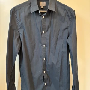 Mörkblå slim fit skjorta från H&M - Stilren mörkblå skjorta från H&M i slim fit-modell. Skjortan har klassisk krage, vita knappar framtill och långa ärmar.