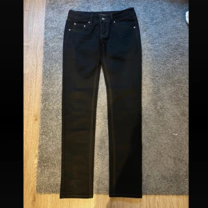 Svarta Nudie Jeans, storlek 28/32 - Snygga svarta jeans från Nudie Jeans med klassisk femficksdesign och kontrastsömmar i orange. Modellen har rak passform och normal midja, med Nudie-logga på bakfickan och läderpatch i midjan. Perfekt för dig som gillar stilrena och tidlösa jeans.