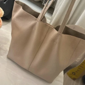 Beige shoppingväska från Rizzo - Snygg och rymlig shoppingväska från Rizzo i beige skinn med mjuk, lite grov struktur. Väskan har två långa handtag och stängs med magnetknapp. Insidan är fodrad i mocka och har en enkel, stilren design utan extra fack. Perfekt för dig som gillar minimalistisk stil.