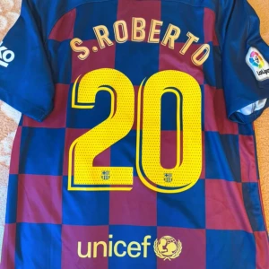 FC Barcelona S. Roberto matchtröja Nike - FC Barcelona matchtröja med S. Roberto och nummer 20 på ryggen. Tröjan är kortärmad, har blått och rött rutigt mönster, officiella loggor från La Liga, Rakuten och UNICEF samt klubbmärke på bröstet. Tillverkad i lätt och ventilerande polyester.