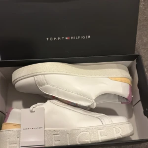 Vita sneakers Tommy Hilfiger strl 40 - Snygga vita sneakers från Tommy Hilfiger med rund tå och platt sula. Skorna har färgglada elastiska band i pastellfärger över vristen och detaljer i rosa och gult på hälen. Perfekta för dig som gillar stilrena men lekfulla detaljer.