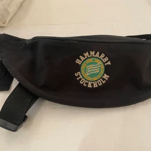Svart Hammarby magväska med broderi - Svart magväska med Hammarby Stockholm-broderi i grönt och gult framtill. Väskan har justerbart band och dragkedja upptill. Perfekt för att bära med sig det viktigaste och visa sitt lagstöd. Enkel och sportig design. 