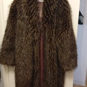 Brun lång pälsjacka med fluffig look - Säljer en riktigt fet lång pälsjacka i mörkbrun ton med fluffig och voluminös känsla. Jackan har bred krage och långa ärmar, perfekt för dig som vill sticka ut och hålla dig varm. Den har en loose passform och ger en cool vintagevibe.