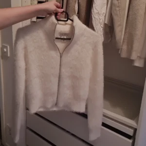 Creme/beige fluffig kofta från Ruby Moon - Supermjuk och fluffig creme kofta från Ruby Moon med dragkedja framtill. Croppad modell med lång ärm och hög krage, perfekt för att ge din outfit en cozy vibe. Materialet är stickat och har en lyxigt lurvig känsla som gör den extra mysig.