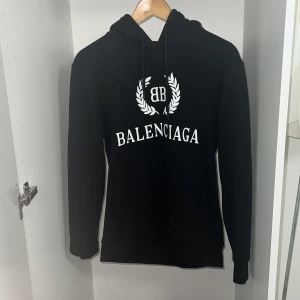 Svart Balenciaga hoodie med tryck - Svart hoodie från Balenciaga med vit logotyp och BB-emblem på bröstet. Tröjan har huva med snörning, känguruficka och långa ärmar. Perfekt nu till vinter och för dig som gillar streetwear och exklusiva märken. Äkta. Storlek M men skulle säga att den passar dig som är i storlek S. För fler bilder skriv