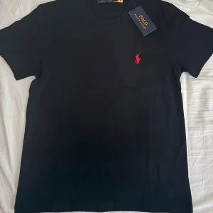 Svart t-shirt från Polo Ralphe Lauren - Svart t-shirt från Polo RalpheLauren med klassisk rund hals och korta ärmar. T-shirten har det ikoniska röda broderade Polo-logot på bröstet. Tillverkad i mjuk bomull för en skön och stilren look.