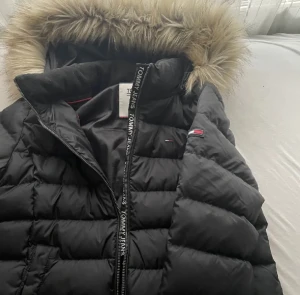 Svart dunjacka Tommy Jeans med päls - Svart dunjacka från Tommy Jeans med huva och snygg fuskpäls. Jackan har quiltad design, dragkedja med logotypdetaljer och två sidofickor. Perfekt för kalla dagar och har en modern, stilren look som funkar till många outfits. Helt oanvänd och köpt för 2599, villig att gå ner i pris vid snabb affär🤍