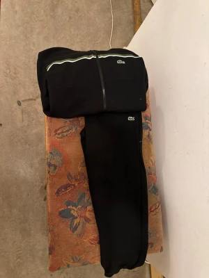 Svarta mjukisbyxor från Lacoste L - Svarta mjukisbyxor från Lacoste med elastisk midja och snörning. Byxorna har gröna och vita ränder längs sidorna och klassisk Lacoste-logga på benet. Tillverkade i mjukt bomullsmaterial för maximal komfort och avslappnad stil. Storlek : L , OBS! Pris kan diskuteras vid snabb affär