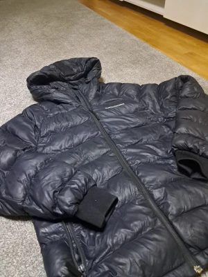 Svart dunjacka Peak Performance - Säljer en svart dunjacka från Peak Performance med huva och dragkedja. Jackan har quiltad design, ribbade muddar och logga broderad på bröstet samt ärmen. Perfekt för kalla dagar och riktigt snygg passform.