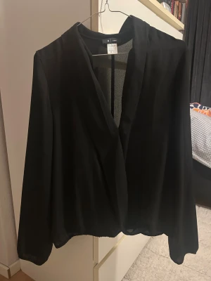 Svart omlottblus från La Redoute - Säljer en svart omlottblus från La Redoute. Blusen har lång ärm, v-ringad omlottfront och är tillverkad i ett lätt, flowy material som ger en snygg siluett. Perfekt för dig som gillar stilrena och enkla plagg med en twist.