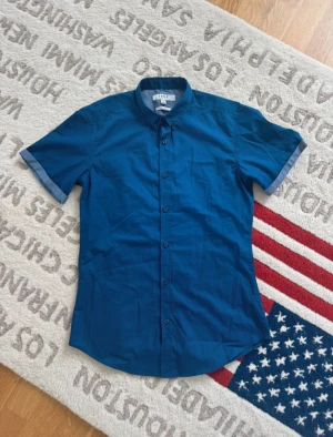 Blå kortärmad skjorta från SMOG - Snygg blå skjorta från SMOG i slim fit-modell. Skjortan har korta ärmar med kontrastfärg på insidan, klassisk krage och knappar hela vägen framtill. Tillverkad i mjuk bomull som är lätt att sköta. Perfekt för dig som gillar stilrena och enkla plagg.