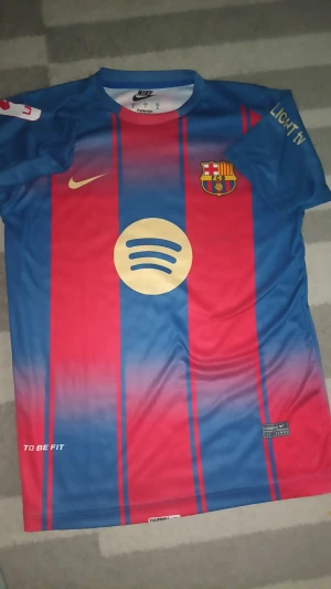FC Barcelona Lamine Yamal #10 Nike tröja - Säljer en FC Barcelona matchtröja med Lamine Yamal #10 på ryggen. Tröjan är kortärmad och har klassiska blå och röda vertikala ränder, med guldfärgade detaljer och loggor. Materialet är lätt och andas, perfekt för fotboll eller häng. Spotify och UNHCR tryck på fram- och baksida.