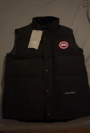 Svart dunväst från Canada Goose - Svart dunväst från Canada Goose med klassisk logga på bröstet. Västen är helt ny och kommer med taggen. Säljes pga att det inte passar min stil och har tappat bort kvittot. Västen är i storlek S