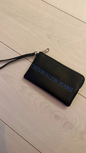 Svart plånbok Calvin Klein Jeans - Snygg svart plånbok från Calvin Klein Jeans med blå logga framtill. Tillverkad i slätt skinnliknande material och har dragkedja samt handledsrem för enkel användning. Perfekt för kort, kontanter och småprylar.