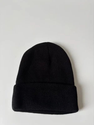 Svart klassisk stickad mössa - En stilren svart mössa med uppvikt kant, perfekt för kalla dagar. Enkel design som funkar till alla outfits och ger en cool vibe. Stickad i mjukt material som sitter skönt på huvudet. Helt ny