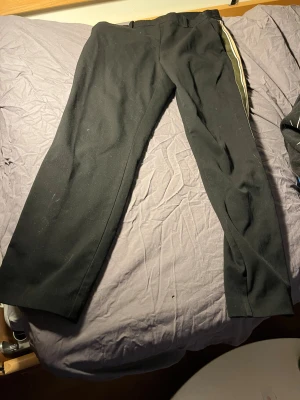 Svarta kostymbyxor med sidoremsa - Snygga svarta kostymbyxor med en vit och olivgrön sidoremsa längs benet. Byxorna har rak passform och klassiska bälteshällor. Perfekt för dig som vill ha en stilren look med en twist. Materialet känns mjukt och har en lätt stretch.