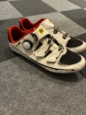 Mavic vita cykelskor med röd insida - Säljer ett par Mavic cykelskor i vitt med svarta och röda detaljer. Skorna har en rund tå, ventilerande meshpartier, justerbar snörning med BOA-system och kardborreband. Insidan är röd och sulan är platt för optimal kraftöverföring. Perfekt för dig som vill maxa din cykling.
